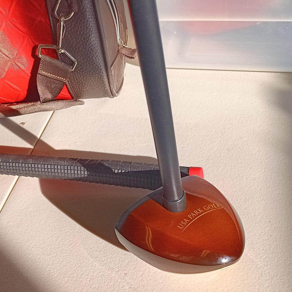 Close-up view of a USA Park Golf club head resting on a light beige surface. The club head is dark reddish-brown wood with the words "USA PARK GOLF" engraved in gold lettering. A dark grey shaft extends upward, and a portion of a black grip is visible in the background. A brown leather golf bag is visible in the upper left corner.